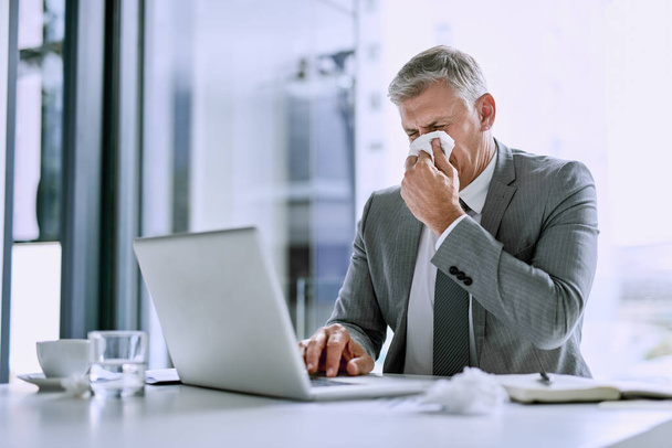 Может, мне стоит поискать информацию об этом гриппе. больной бизнесмен сморкается и работает над ноутбуком в офисе - Фото, изображение