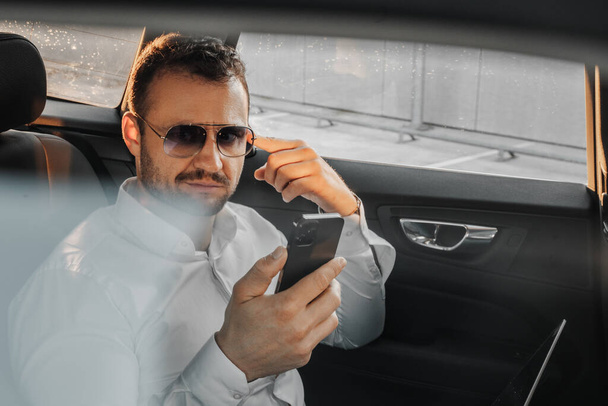 Shot of serious businessman with sunglasses and mobile phone sitting inside expensive car. - Foto, imagen