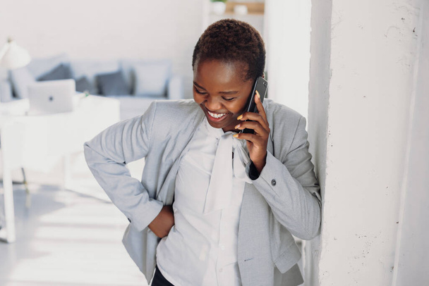 Young African American businesswoman standing in the modern office, and talking on mobile phone with the client. Modern technology. Smiling employee using - Foto, Imagem