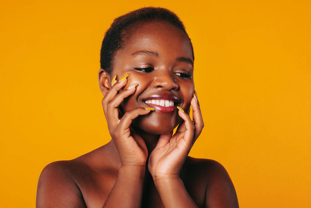 Όμορφη γυναίκα από την Αφρική απολαμβάνοντας απαλότητα του καθαρού προσώπου μετά από τη διαδικασία φροντίδας του δέρματος στο σαλόνι σπα, απομονώνονται σε πορτοκαλί φόντο. Αισθητική δερματολογία έννοια - Φωτογραφία, εικόνα