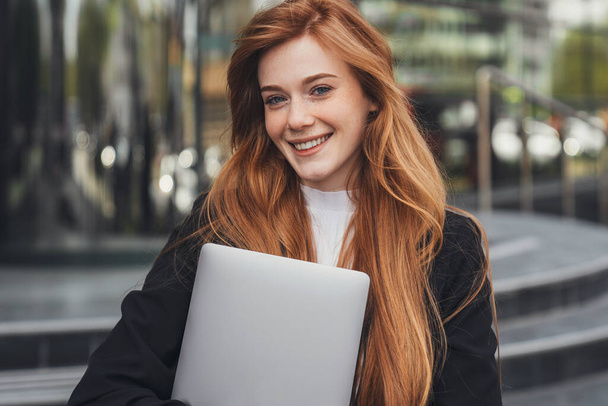 Portrait of smiling read-headed businesswoman standing at office building holding her laptop and looking at camera. Woman beauty face. Business portrait. - Foto, afbeelding