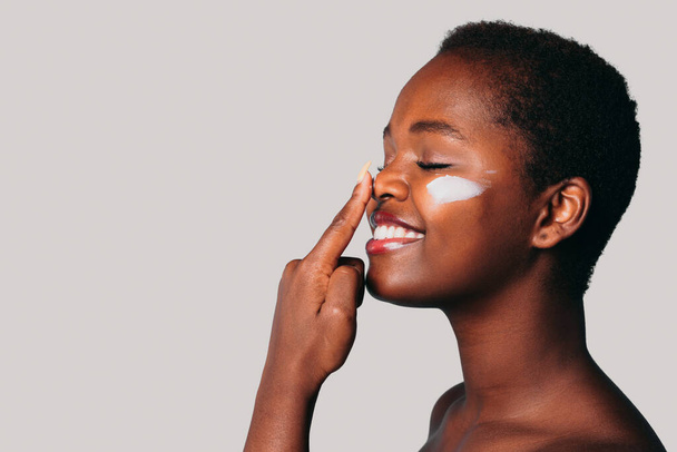 Free space for text. Profile view of an African woman touching her nose with her index finger applying cream on her face, isolated over gray background with - Photo, image