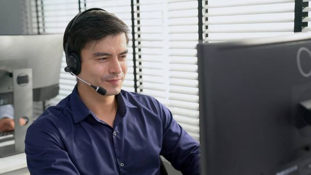 Young competent asian male call center agent working at his computer while simultaneously speaking with customers. Concept of an operator, customer service agent working in the office with headset. - Fotó, kép