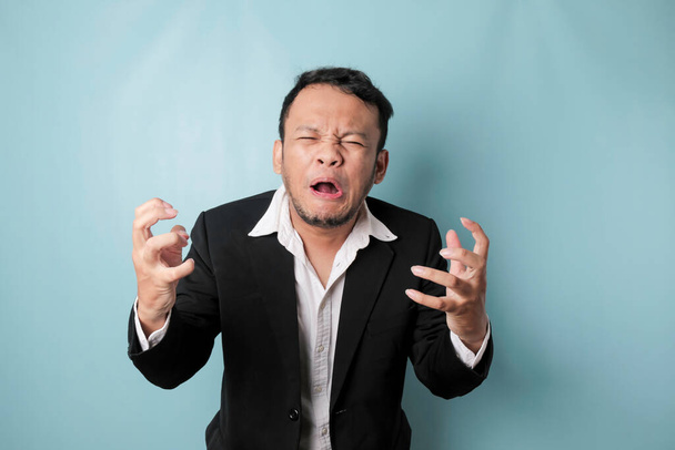 Angry and mad face of Asian businessman in black suit on isolated blue background. - Photo, image