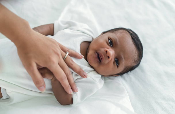 Yatakta uzanmış, yatak odasındaki kameraya bakan sevimli Afro-Amerikan bebek portresi. Anne küçük çocuğu uyutuyor. Çocuk bakımı, siyahi aile, yeni doğan erkek bebek. Yeni hayat - Fotoğraf, Görsel