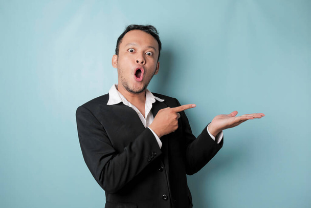 Shocked Asian man wearing suit pointing at the copy space beside him and smiling, isolated by blue background - Photo, image