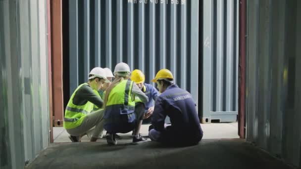 Group of multiracial workers in safety uniforms and hardhats sit and discuss logistics with White male manager, trading shipping goods for cargo transportation industry, shot from inside a container.  - Metraje, vídeo
