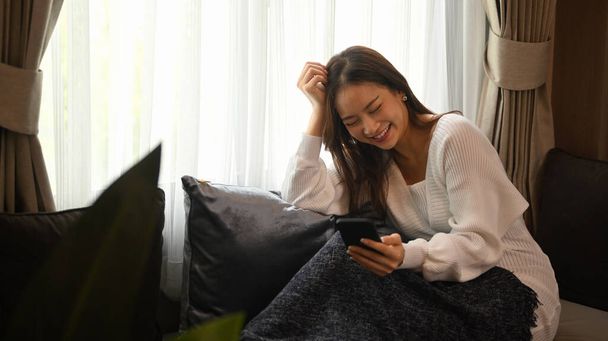 Smiling young woman relaxing on comfortable couch and enjoying chatting online with her friends via smart phone. - Φωτογραφία, εικόνα