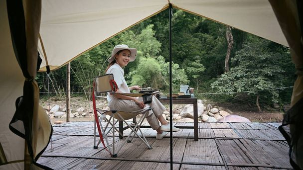 Peaceful asian woman sitting on folding chair near river bank during camping in nature park. Travel, adventure and vacation concept. - Foto, immagini