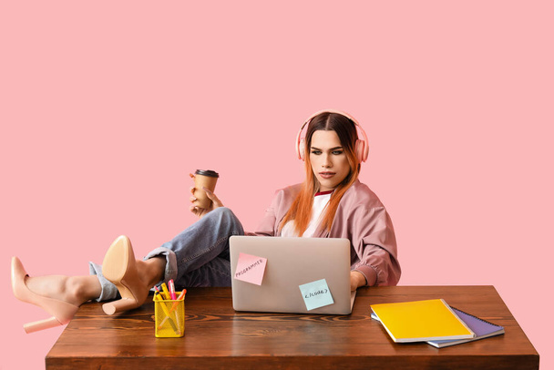 Трансгендерний програміст з ноутбуком за столом на рожевому фоні - Фото, зображення