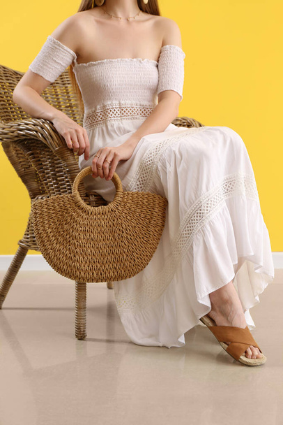 Elegant woman sitting in chair and holding stylish rattan handbag on yellow background - 写真・画像