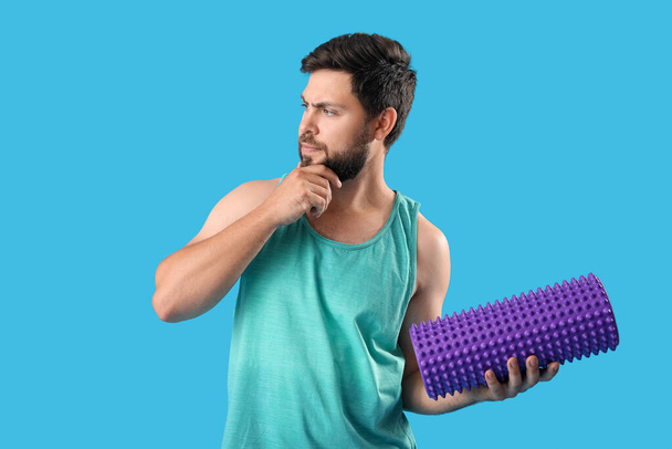 Thoughtful young man with foam roller on blue background - Foto, afbeelding