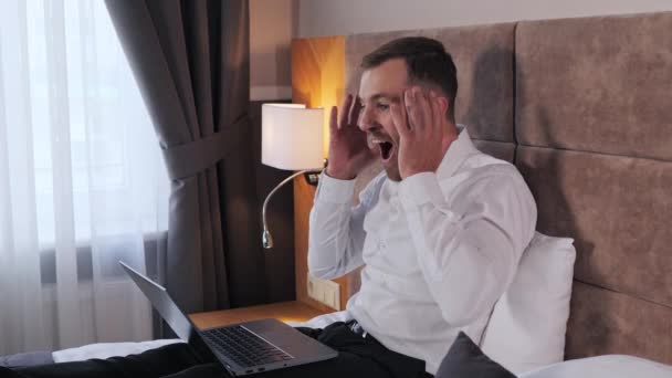Happy business man sitting on the bed working on laptop in hotel room celebrating success, amazing incredible news, gesturing hand, shouting yes winner. Financial stock sports betting crypto exchange - Footage, Video