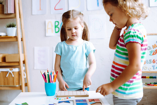 Две сконцентрированные милые маленькие девочки-ученицы, стоящие за белым столом с цветными карандашами, играют в настольные игры в ярком классе. Ремесла, раннее развитие, дошкольное образование, форвардность. - Фото, изображение
