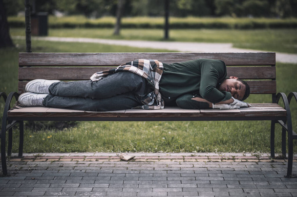 Social problems. African american young man lying sleeping on bench in city park during day - Foto, Imagem