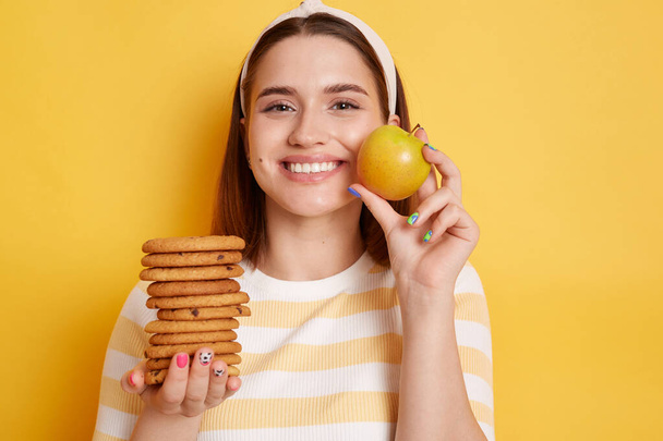Attractive slip happy smiling woman wearing striped shirt and hair band, holding cookies and apple, prefer healthy eating instead of junk food, posing isolated over yellow background. - 写真・画像