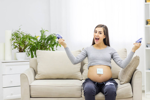 Photo of pregnant woman holding a paper with the sex of her future child on her stomach and childs shoes in her hands. Concept of pregnant woman. - Fotoğraf, Görsel