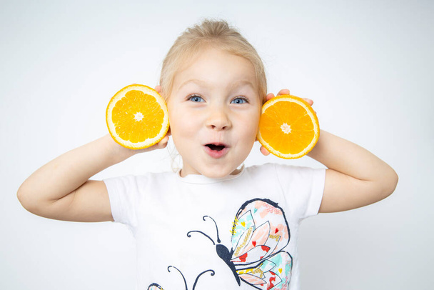 Surprised child girl blonde holding two cut halves of an orange on a white background. - 写真・画像