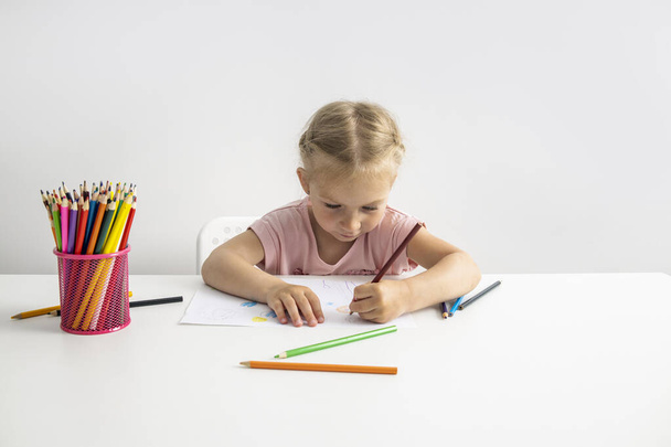 Блондинка девочка рисует цветными карандашами сидя за столом. - Фото, изображение