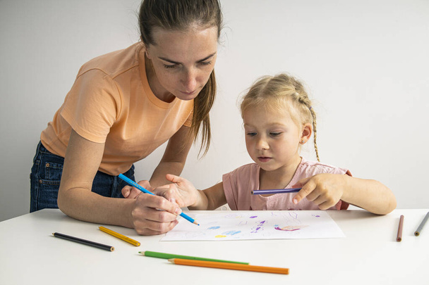 Mom helps her daughter draw with colored pencils on paper at the table. - Fotoğraf, Görsel