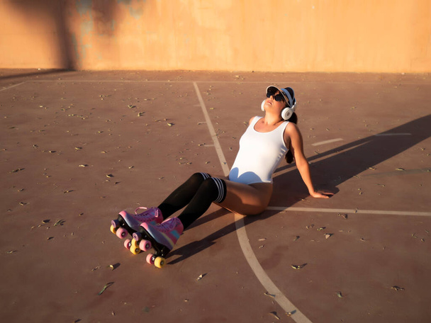 From above relaxed young female traveler in swimwear and roller skates leaning back and listening to music while sitting on sunlit sports ground - Foto, Imagen