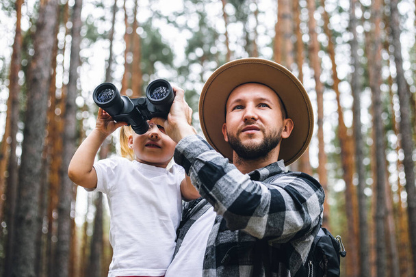 森の中をハイキングしながら、帽子とバックパックの男と子供は双眼鏡を通して見ています。山や森への家族のハイキング. - 写真・画像