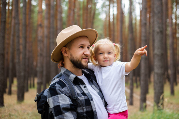 Чоловік у капелюсі тримає дитину на руках, ходячи в ліс. Сімейний похід в гори або ліс
. - Фото, зображення