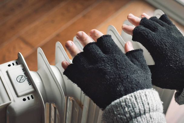 Κοντινό πλάνο των χεριών ενός ατόμου φορώντας μια μάλλινη ζακέτα και μαύρα γούνινα γάντια χωρίς δάχτυλα κοντά σε ένα οικιακό καλοριφέρ προσπαθεί να κρατήσει ζεστό το χειμώνα - Φωτογραφία, εικόνα