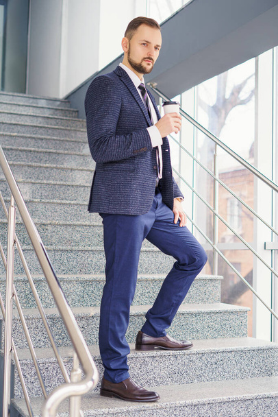 A young man in a business suit on the stairs with a glass of coffee. A businessman on the stairs. - Photo, Image