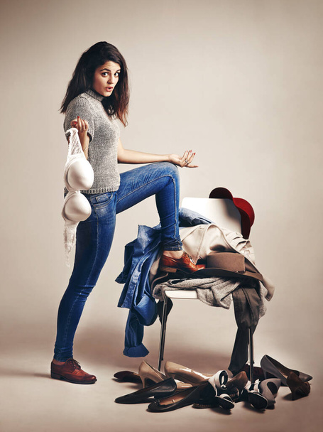 Ακόμα και οι fashionistas έχουν ρεπό. Στούντιο πλάνο μιας νεαρής γυναίκας που διαλέγει ρούχα στοιβαγμένα σε μια καρέκλα πάνω σε καφέ φόντο - Φωτογραφία, εικόνα