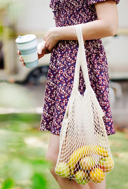 Young female in dress holding reusable silicone foldable drinks mug. eco-friendly. fresh fruits in a beige knitted string bag. Zero waste, healthy food concept. - Zdjęcie, obraz