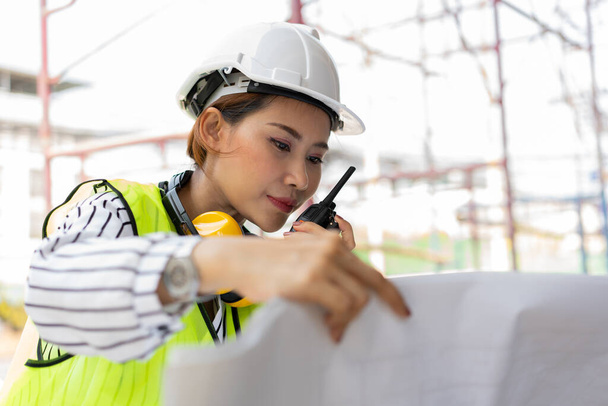 Asian engineer or Young woman Architect put on a helmet for safety and look at Blueprint for Inspect Building factory Construction Site and use walkie talkie for a talk with a contractor. - Фото, изображение