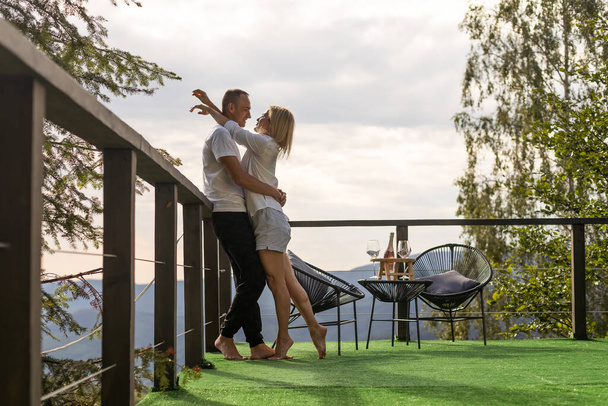 Horizontal sideview shot of a young couple in summer outfit enjoying the mountain view from terrace. Copy space - Zdjęcie, obraz
