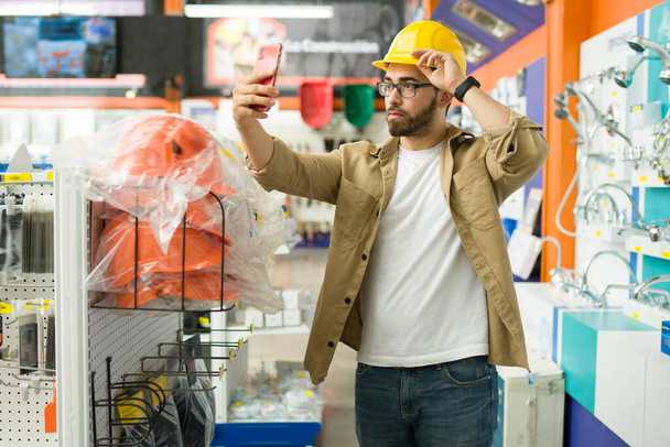 Νεαρός άνδρας ή μηχανικός λαμβάνοντας μια selfie με το smartphone του, ενώ φοράει ένα κίτρινο κράνος ασφαλείας για να εργαστούν στις κατασκευές - Φωτογραφία, εικόνα