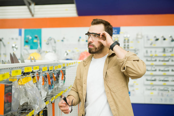 Attractive caucasian man putting on safety glasses and buying gear at the hardware store for his construction work - Foto, afbeelding