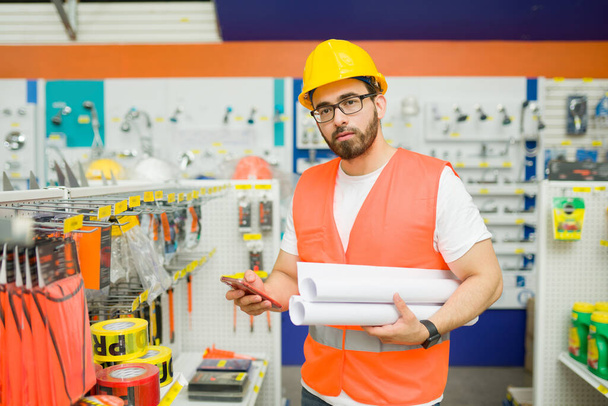 Красивый мужчина-инженер с защитным шлемом и жилетом, держащий чертежи во время покупок в хозяйственном магазине - Фото, изображение