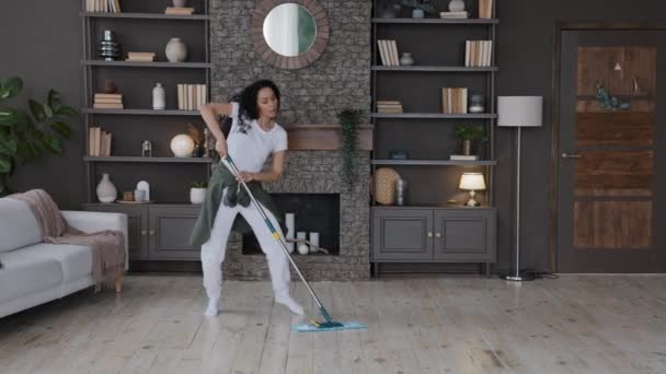 Молодая энергичная женщина домохозяйка чистит дома моющие полы в гостиной, танцуя со шваброй счастливая активная жизнерадостная латиноамериканка танцует под музыку танцовщица, исполняющая хореографию наслаждаясь уборкой - Кадры, видео