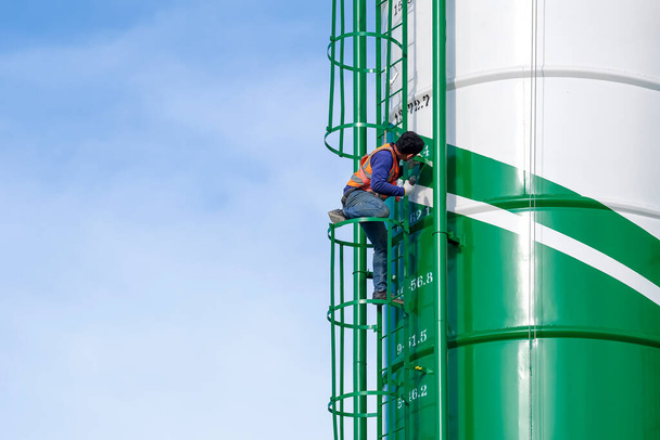 Maintenance worker with safety equipment is repairing steel ladder  on concrete storage silo against blue sky background - Photo, Image