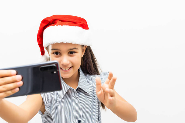 маленькая девочка в шляпе Санты со смартфоном на белом фоне. - Фото, изображение