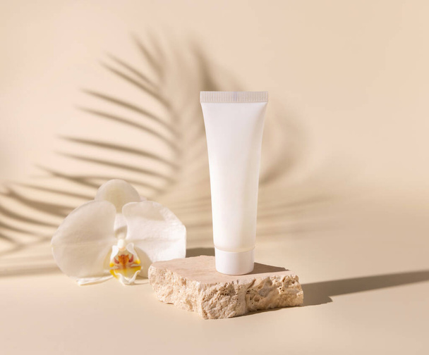 Λευκό πλαστικό σωλήνα κρέμα σε τραβερτίνη πέτρα κοντά σε λευκό λουλούδι ορχιδέα σε ανοιχτό μπεζ, Palm φύλλα σκληρές σκιές, κοντά, mockup. Προϊόν ομορφιάς Skincare. Φυσικά καλλυντικά. Τροπική λιπασματοποίηση - Φωτογραφία, εικόνα