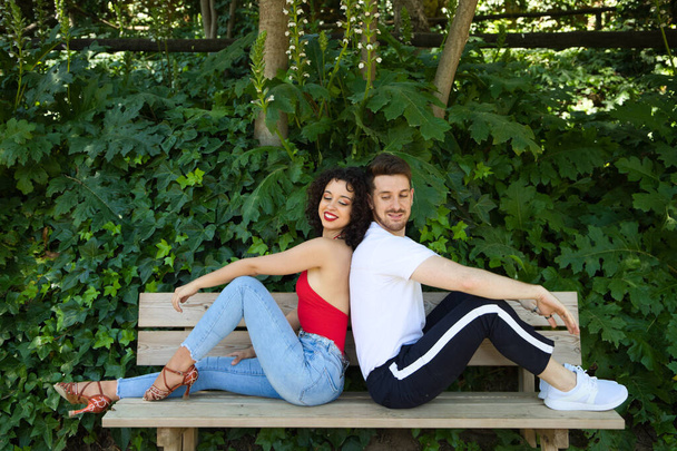 Egy jóképű fiatalember és egy gyönyörű nő ülnek a fa padon háttal egymásnak. Mindegyik más-más oldalra néz, ahogy boldogan néznek egymásra a szemük sarkából.. - Fotó, kép
