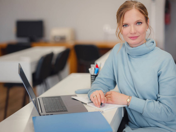 Ofiste çalışan mavi elbiseli bir iş kadını. Çevrimiçi iş, iş yerinde genç bir profesyonel. Masada dizüstü bilgisayar kullanarak. çevrimiçi webinar izlemeyi öğrenmek - Fotoğraf, Görsel
