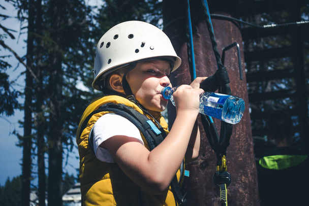 Small boy drinking water while taking a break on treetop during canopy tour in nature.  - Zdjęcie, obraz