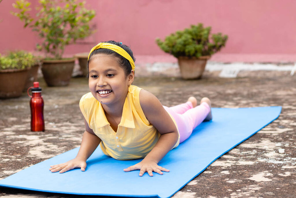 Индийская девочка практикующая йогу в улыбающемся лице на коврике для йоги на открытом воздухе - Фото, изображение