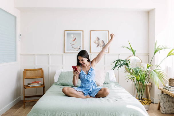Happy young woman celebrating good news on mobile phone. Excited female cheering online success on cellphone sitting on bed - Foto, Bild