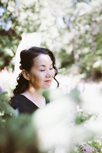Beautiful happy middle-aged Japanese woman in black casual dress summer park, smiling. Eyes closed. Close-up portrait. Sunlight, copy space - Fotó, kép