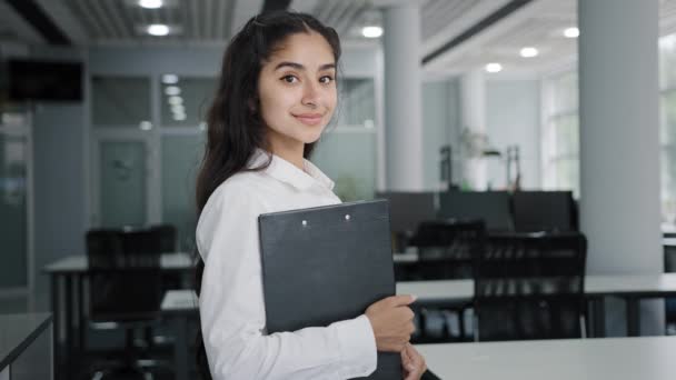 Young confident Indian businesswoman model posing indoors smiling female manager office worker trainee stands holding folder with documents in hand looking at camera advertises business corporation - Imágenes, Vídeo