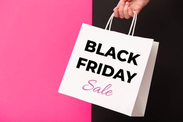 Female hand holding white shopping paper bag with Black Friday Sale text on pink and black background. Sale, discount, shopping concept - Zdjęcie, obraz