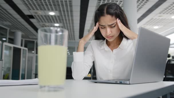 Young tired woman manager sits in office feels headache suffers from pain overwork unhealthy girl office worker feeling unwell migraine pressure syndrome focus on glass of pain medication treatment - Footage, Video