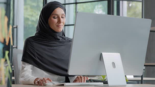 Confident arab businesswoman manager in hijab sitting in modern office working on internet looking at monitor typing on keyboard successful business lady creates project using computer technology - Filmati, video